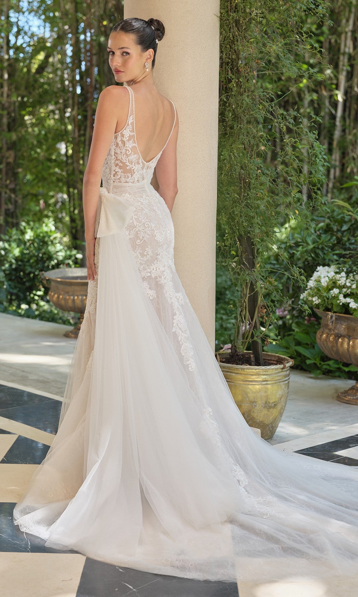 A woman in the Andrea and Leo WL068 long wedding dress with a sheer corset bodice and long train stands outdoors on a checkered floor, looking over her shoulder. Lush greenery and large planters are in the background.